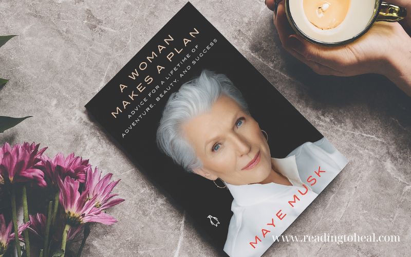 The book 'A Woman Makes a Plan' by Maye Musk placed on a marble surface next to a cup of coffee and pink flowers.