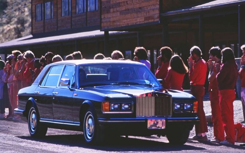 Osho sitting inside a luxury dark blue Rolls Royce surrounded by followers in red robes at Rajneeshpuram.