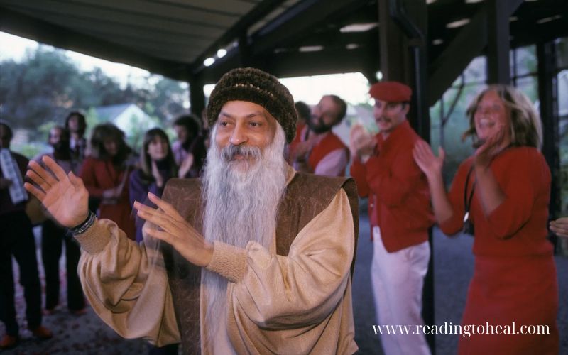 Osho smiling and gesturing toward a group of followers in red clothing